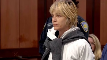 Mujer de cabello rubio corto con sudadera blanca y un abrigo gris sobre los hombros junto a un agente de policía
