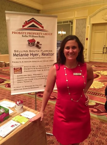 Una mujer sonriente, Melanie Lauren Hyer, con un vestido rojo y un collar, posa junto a un cartel de 'Probate Property Group' en un evento
