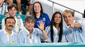 Seis personas sentadas en gradas de estadio. Cinco llevan camisetas de fútbol de Argentina con franjas celestes y blancas. Una mujer saluda, un hombre rubio señala.