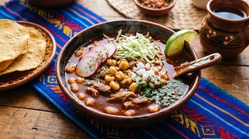 Plato de barro con pozole rojo, rábanos, col, cebolla y un trozo de lima, junto a tostadas sobre un mantel azul.