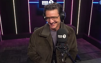 Pedro Pascal sonriendo y riendo, con gafas y auriculares negros, en un estudio de radio. Un micrófono de BBC Radio 1 está en primer plano. Luces de neón en la pared