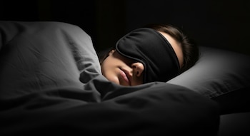 Primer plano de una persona durmiendo de lado, usando un antifaz negro, cubierta por ropa de cama gris y con el rostro parcialmente visible en la oscuridad.