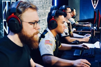 Cuatro hombres sentados frente a monitores de computadora con auriculares. Se aprecian teclados, ratones y un controlador. Uno de los hombres lleva la bandera de Estados Unidos