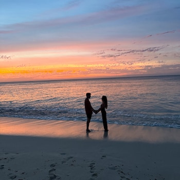 Siluetas de un hombre y una mujer descalzos en la orilla de una playa, tomados de la mano, con el mar y un cielo anaranjado al atardecer