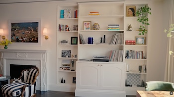 Una sala de estar con chimenea blanca, un cuadro de playa, un sillón a rayas y una estantería empotrada llena de libros, discos y objetos
