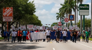 Vista frontal de decenas de personas marchando en una calle urbana soleada. Llevan pancartas y hay tiendas como Home Depot, Trader Joe's, Target y Aldi a los lados.