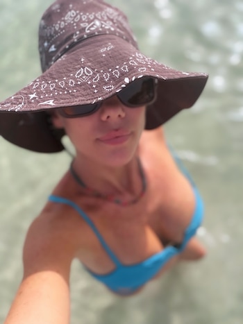 Mujer con sombrero de tela estampado, gafas de sol y bikini azul en el agua, con collar en el cuello, vista desde arriba