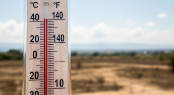 Un termómetro blanco muestra una temperatura alta, con el mercurio rojo marcando casi 40°C y más de 100°F, frente a un paisaje árido borroso.