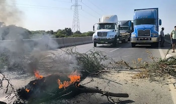 Vista de una carretera bloqueada por ramas en llamas y humo denso, con varios camiones y personas detenidos en el asfalto bajo un cielo claro