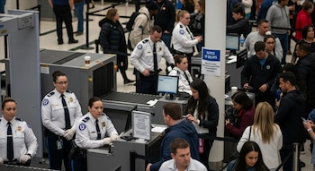 Vista aérea de un control de seguridad de aeropuerto abarrotado con agentes de la TSA uniformados y numerosos viajeros haciendo fila para pasar la inspección.
