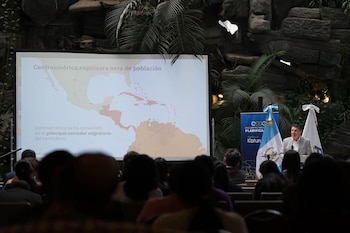 Un hombre habla en un podio junto a la bandera de Guatemala ante una pantalla con un mapa del continente americano sobre migración