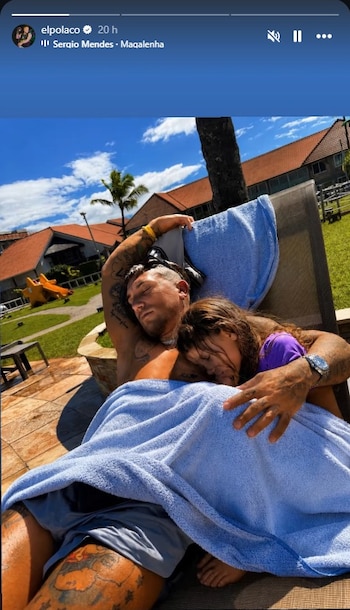 Hombre tatuado y niña pequeña durmiendo en reposera. Ambos cubiertos con toalla celeste. Fondo con palmeras, césped y edificios de techos rojizos