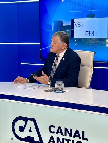 Hombre de negocios de cabello gris, vestido con traje azul, sentado en un escritorio blanco con un teléfono, un vaso de agua y papeles, frente a una pantalla azul
