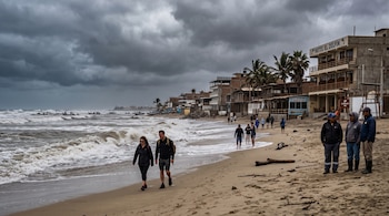 Personas caminan por una playa de arena junto a un mar con oleaje blanco, frente a varios edificios y bajo un cielo de nubes grises.