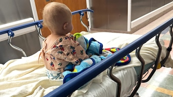 Un bebé con bata de hospital y pañal, cubierto con una manta azul, sentado de espaldas en un transportín médico con barandillas azules en un hospital