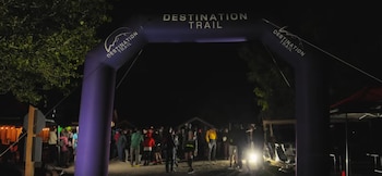 Un arco inflable morado con la frase Destination Trail se alza de noche sobre un grupo de personas reunidas en un terreno de tierra
