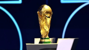 Primer plano del Trofeo de la Copa Mundial de la FIFA, dorado con una base verde, exhibido en un pedestal blanco contra un fondo oscuro con luces azules