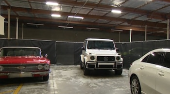 Vista frontal de un coche clásico rojo, un SUV blanco Mercedes-Benz G-Class y la parte trasera de un sedán blanco, estacionados en un almacén