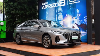 Sedán Chery Arrizo 8 plateado exhibido en una plataforma de madera. Detrás, un cartel azul muestra el nombre del modelo y un paisaje urbano. Un cargador eléctrico verde a la izquierda