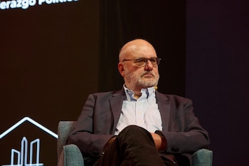 Hombre con anteojos, barba canosa y saco oscuro sentado en un sillón gris durante un evento