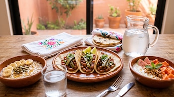 Tacos de frijol con nopales, avena con plátano, fruta fresca, tortillas en servilleta bordada, cubiertos, vaso y jarra de agua sobre mesa de madera.