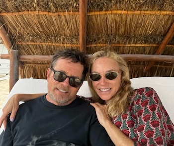 Michael J. Fox y Tracy Pollan posan sonrientes usando gafas de sol, recostados bajo una estructura con techo de paja en un día soleado