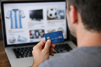 Un hombre sostiene una tarjeta de crédito con una mano frente a la pantalla desenfocada de una computadora portátil que muestra artículos de venta online.