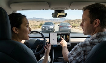 Interior de un coche, mujer al volante y hombre señalando pantalla táctil con video; la carretera y montañas áridas visibles a través del parabrisas