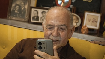 Hombre de edad avanzada con bigote canoso y camisa marrón oscuro sentado en un sofá amarillo, mirando fijamente un teléfono móvil gris. Fotos enmarcadas borrosas al fondo