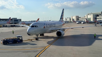 Avión blanco de American Airlines siendo empujado por un tractor azul en la pista de un aeropuerto soleado, con un trabajador y edificios en el fondo
