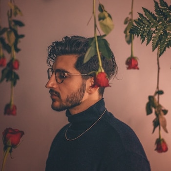 Hombre de perfil con cabello oscuro, barba, gafas, y cuello alto negro. Rosas rojas colgantes y hojas verdes sobre fondo liso