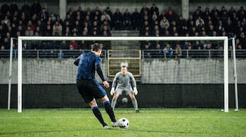Un jugador de fútbol con camiseta azul patea un balón blanco y azul hacia el arco. Un arquero lo espera. Tribunas con público borroso bajo la lluvia.