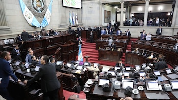 Vista general de la sala de sesiones del Congreso de Guatemala con numerosos diputados sentados en escritorios de madera, frente a pantallas, durante una sesión