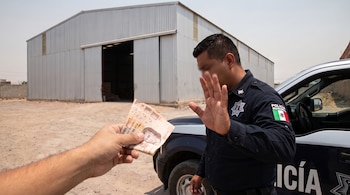 Una mano extiende billetes de 200 pesos mexicanos hacia un oficial de policía que alza una mano en señal de detente, junto a un vehículo policial y un almacén.