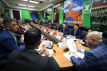Varios hombres con traje formal permanecen sentados alrededor de una mesa de reuniones con micrófonos y letreros con nombres