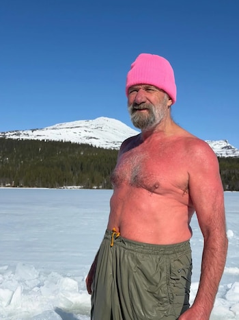 Wim Hof, con piel rojiza y gorro rosa, sin camisa, de pie en un paisaje nevado. Al fondo, un lago helado, árboles y montañas cubiertas de nieve bajo un cielo azul