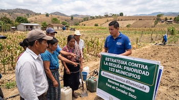 Un técnico con polo azul sostiene un cartel sobre la sequía junto a varios agricultores en un terreno agrícola seco.
