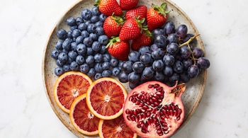 Un plato contiene arándanos, fresas, uvas oscuras, rodajas de cítricos rojos y una granada abierta, sobre una superficie clara.