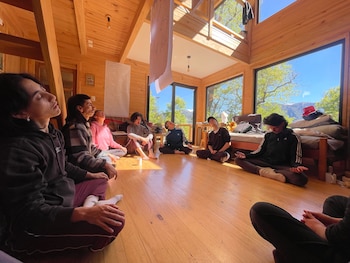 Grupo de personas sentadas en círculo sobre un piso de madera en una habitación luminosa con grandes ventanas. Al fondo se ven árboles verdes y montañas