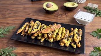Una bandeja negra con bastones dorados y sésamo negro, junto a un recipiente con salsa blanca, medio aguacate y un cartel con texto.