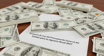 Un documento con el título Proyecto de Ley del Presupuesto General del Estado para 2027 sobre un escritorio de madera, rodeado de billetes de cien dólares.