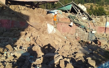 Hombre con casco blanco y chaleco naranja sobre escombros de un edificio derrumbado. Restos de paredes, techo y vigas. Árboles al fondo