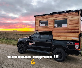 Camioneta pickup negra con camper de madera estacionada en camino de tierra. Fondo de campo y cielo con nubes de atardecer. Texto y emoji
