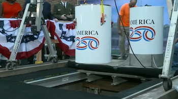 Dos cápsulas cilíndricas blancas con la inscripción "America 250" son bajadas hacia una fosa. Hay varias personas, andamios y banderines patrióticos