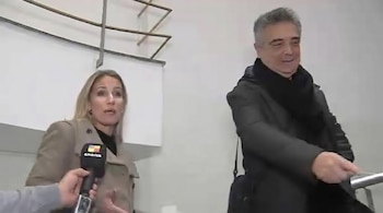 Hombre con cabello gris, abrigo oscuro y bufanda junto a una mujer rubia con abrigo beige, que habla a un micrófono de América.