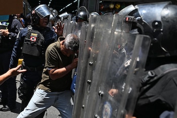 Un hombre de mediana edad con cabello gris, camiseta verde y jeans, empuja con el hombro contra escudos antidisturbios transparentes sostenidos por policías