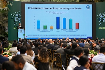 Un orador habla junto a una pantalla con gráficos sobre economía frente a un público sentado en un salón de eventos