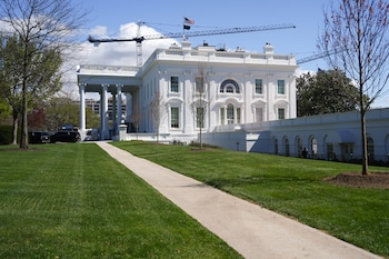 Vista exterior del Ala Este de la Casa Blanca, un edificio blanco con columnas y ventanas arqueadas, con grúas de construcción al fondo, césped verde y un camino