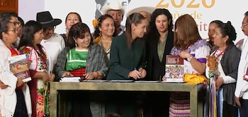 Un grupo de personas, varias con vestimenta tradicional, observa a una mujer que firma documentos en una mesa frente a libros y folletos