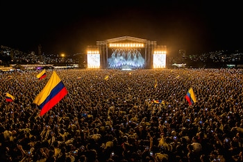 Una multitud observa de noche un escenario iluminado durante un evento musical con banderas de Colombia entre los asistentes.
